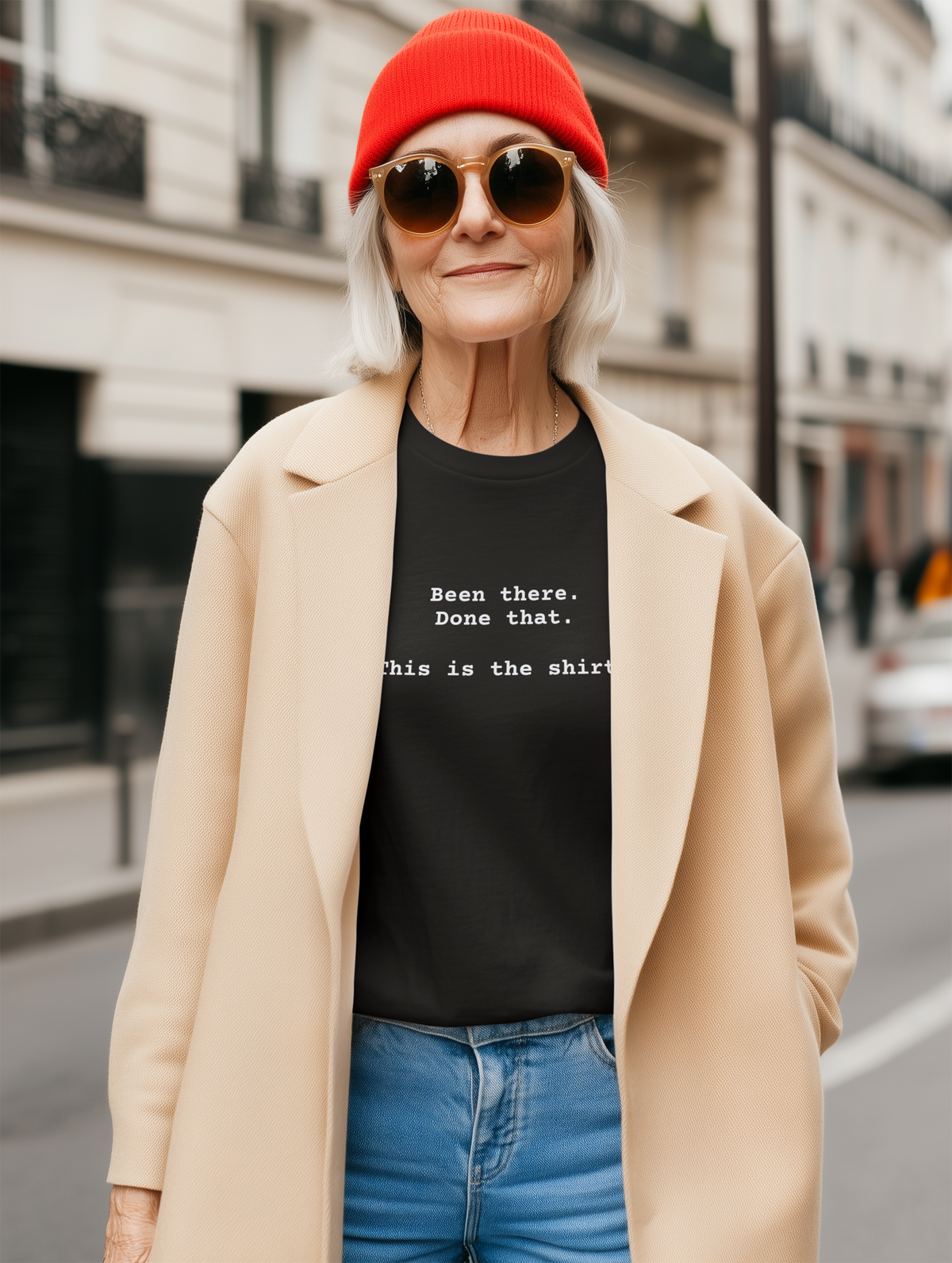 Black t-shirt with text on a white background says "Been There.  Done That.  Here's The T-shirt" in a simple typewriter font. Worn by older woman in red beanie and tan coat.