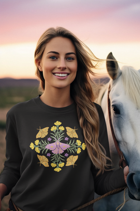 Black long-sleeve crewneck t-shirt featuring a colorful central graphic of a purple butterfly surrounded by yellow flowers and green leaves, with orange butterflies on either side. A mandala. worn by woman holding a horse.
