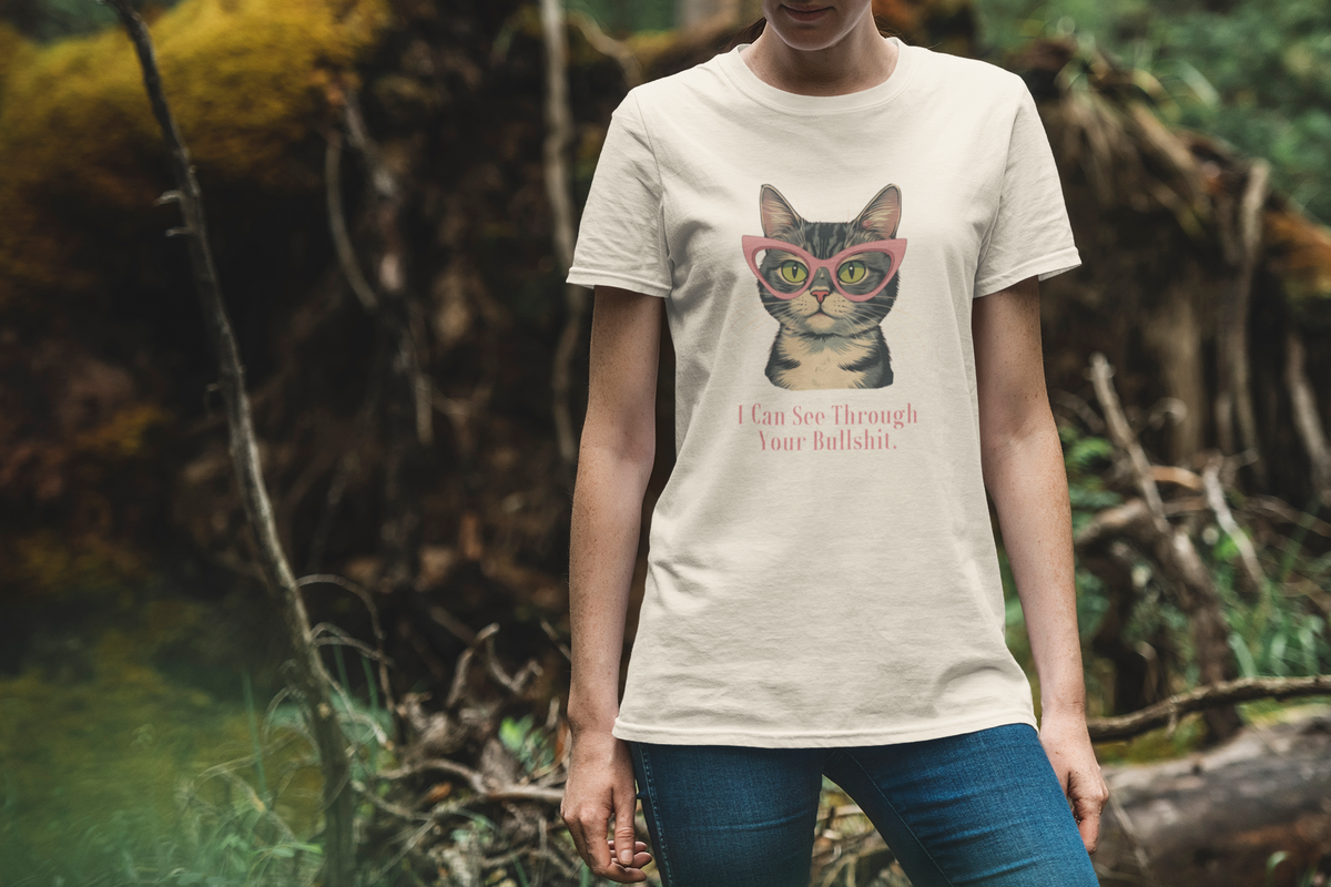 Ivory short-sleeve t-shirt with a graphic of a tabby cat wearing pink glasses and the text 'I Can See Through Your Bullshit.' printed below, displayed on a clean white background. Worn by woman standing near the woods.