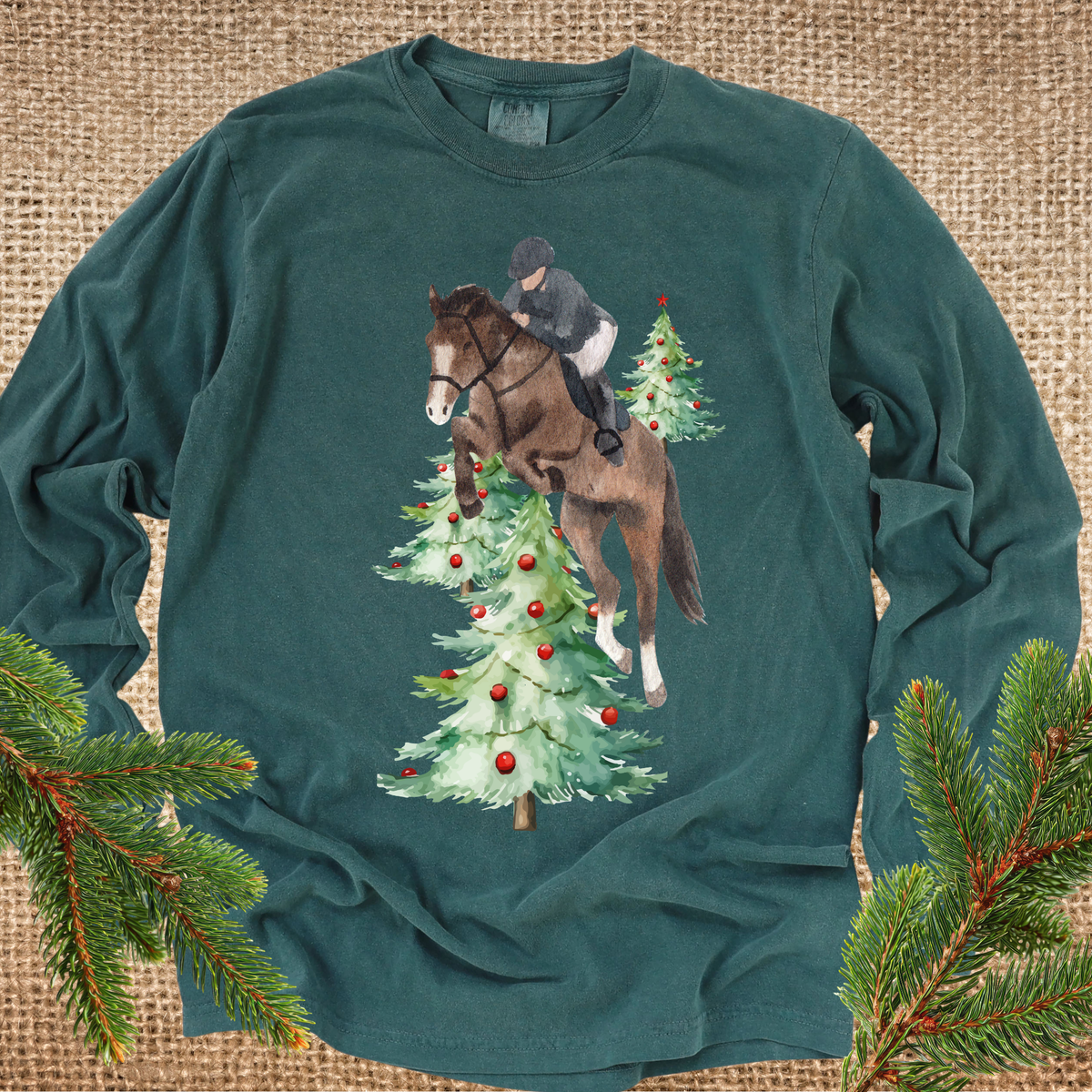 Green  long-sleeve t-shirt with a graphic of a horse and rider jumping over Christmas trees, styled flat on a burlap background with pine branches in the corners.