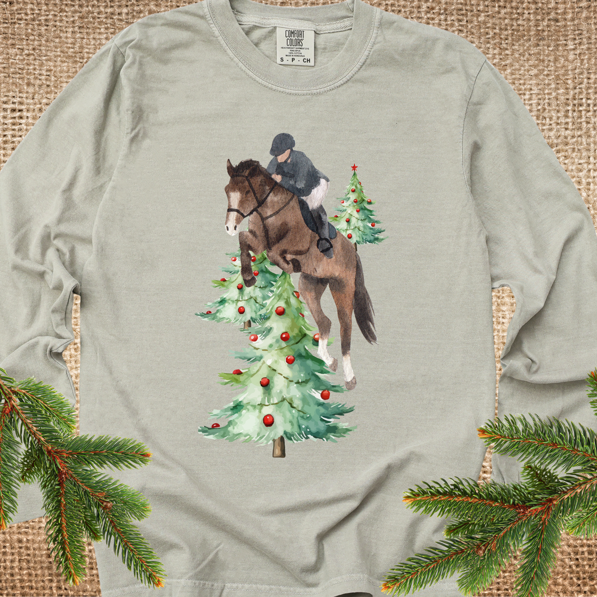 Khaki long-sleeve t-shirt with a graphic of a horse and rider jumping over Christmas trees, styled flat on a burlap background with pine branches in the corners.