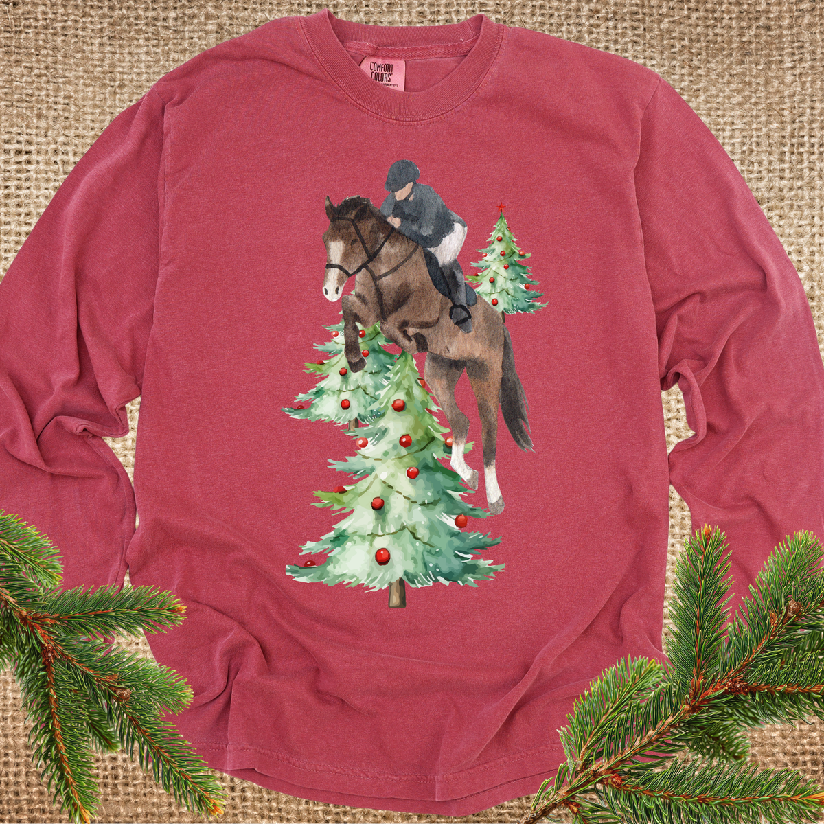 Red long-sleeve t-shirt with a graphic of a horse and rider jumping over Christmas trees, styled flat on a burlap background with pine branches in the corners.