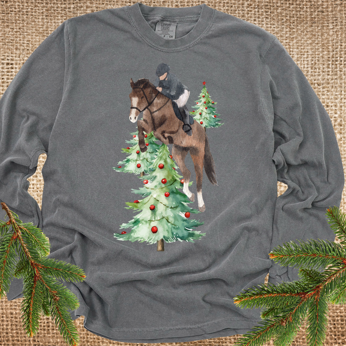 Vintage black long-sleeve t-shirt with a graphic of a horse and rider jumping over Christmas trees, styled flat on a burlap background with pine branches in the corners.