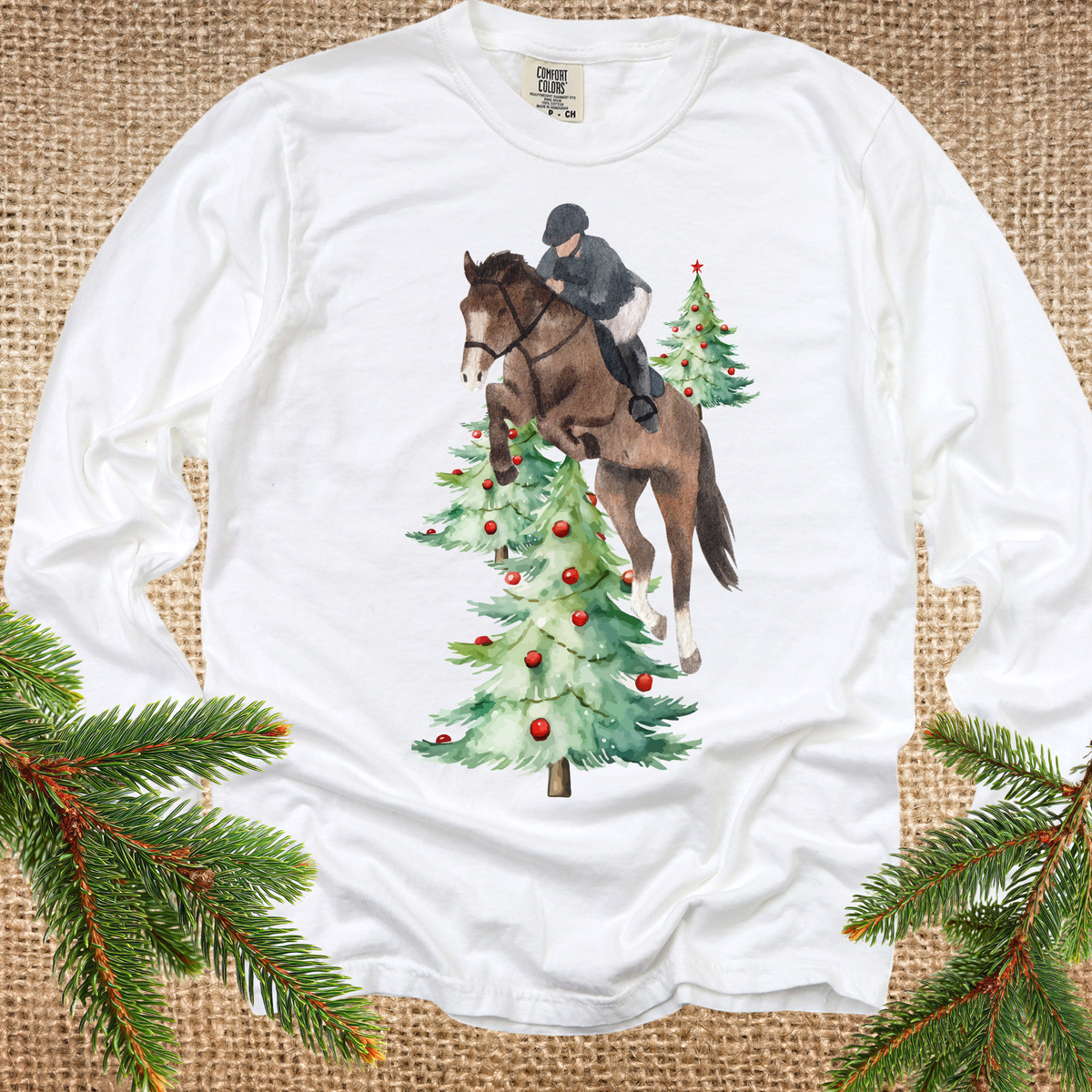 A white long-sleeve t-shirt with a graphic of a horse and rider jumping over Christmas trees, styled flat on a burlap background with pine branches in the corners.