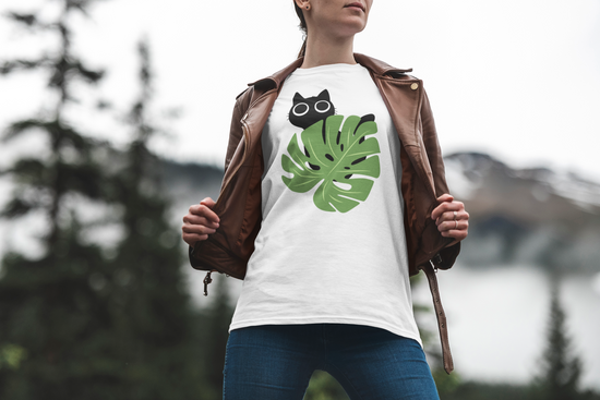 White short-sleeve crewneck t-shirt featuring a graphic of a black cat peeking over a green monstera leaf with white-rimmed glasses, displayed on a clean white background.