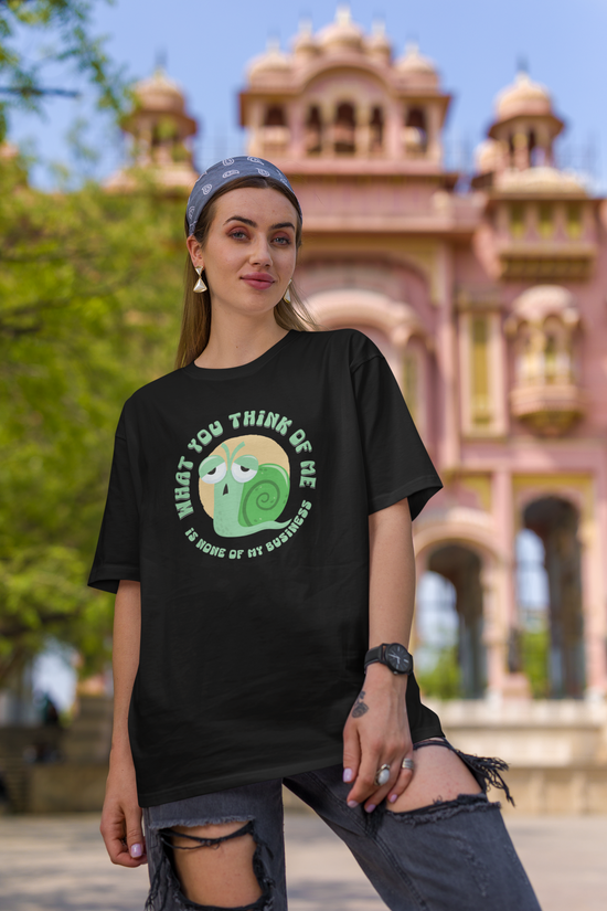 Black short-sleeve crewneck t-shirt featuring a cartoon graphic of a green snail inside a yellow circle, with the text 'WHAT YOU THINK OF ME IS SOME OF MY BUSINESS' printed around it. Worn by woman.